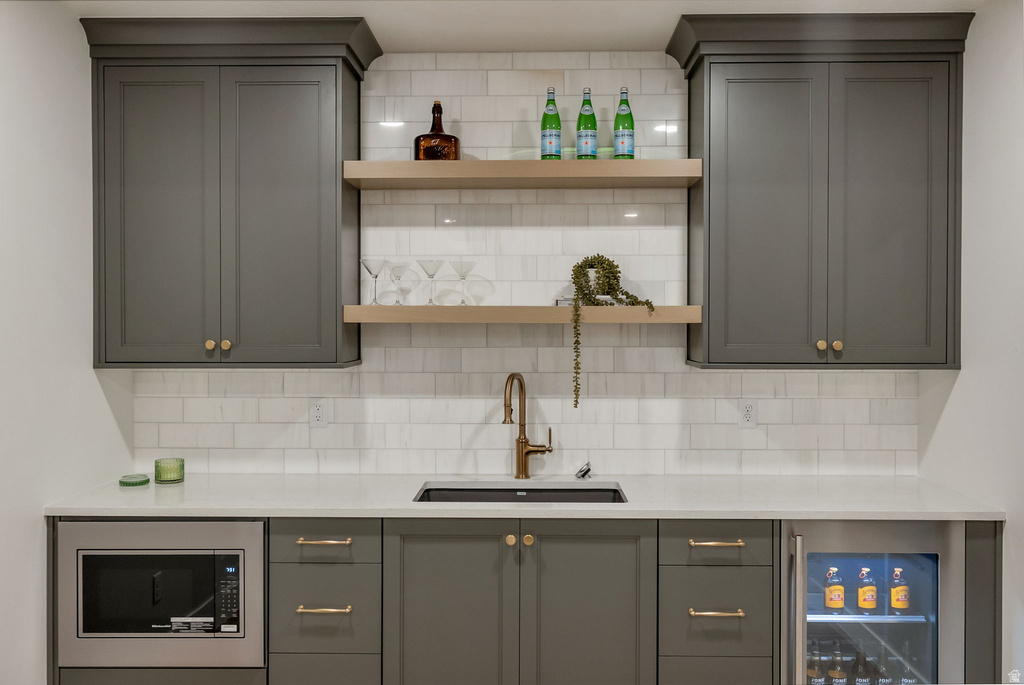 Wet bar featuring gray shaker-style cabinetry, a built-in microwave, under-counter beverage refrigeration, a drop-in sink with a bronze-finish faucet, and open wood-finish shelving against a subway tile backsplash