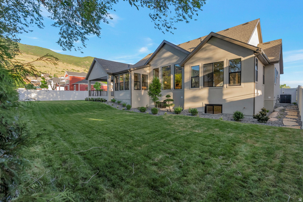 Expansive backyard featuring a manicured lawn, established landscaping, a covered patio with dark railings, a stone pathway, and a stucco exterior with multiple large windows