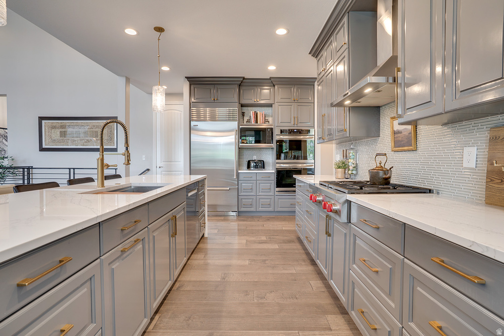Gourmet kitchen featuring extensive gray cabinetry, light wood-finish flooring, white countertops, a large center island with an integrated sink and gold-tone faucet, and stainless steel appliances including a built-in refrigerator and wall ovens