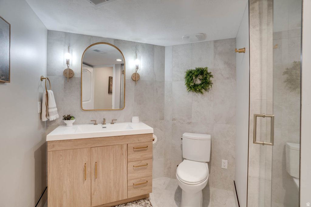 Bathroom featuring a wood-finish vanity with a white countertop and integrated sink, a gold-framed arched mirror, two wall-mounted sconces with clear glass shades, large format grey tile walls, and a frameless glass shower enclosure