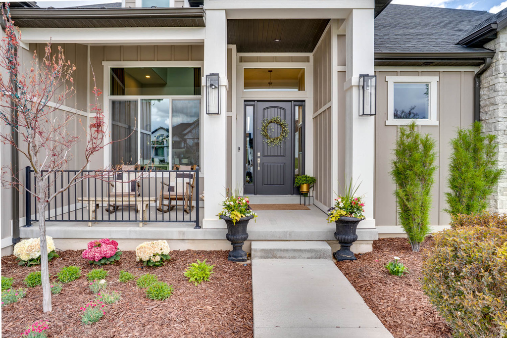 Inviting entryway featuring a dark paneled front door with sidelights, a covered porch with a sliding glass door, board and batten siding, stone veneer accents, and an established landscape with a concrete walkway