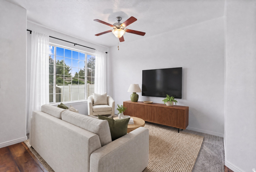Living area featuring a large grid-pattern window, wood-finish flooring, a ceiling fan with integrated lighting, white walls, and a textured area rug