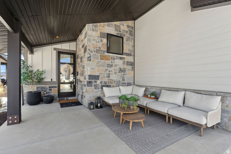 Covered concrete patio with stone veneer accent wall, white horizontal siding, dark wood-finish ceiling, and a modern glass-panel entry door