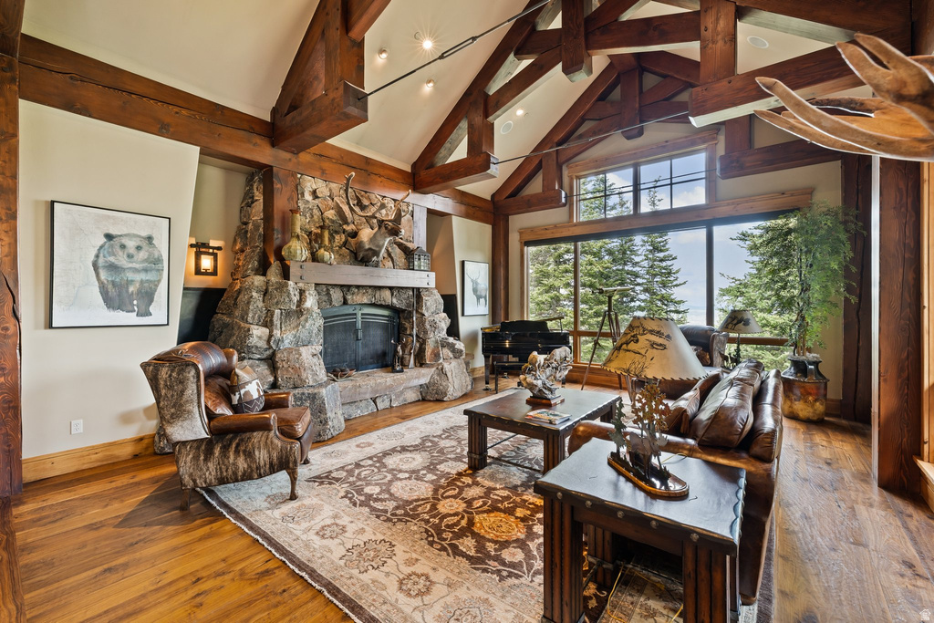 Spacious living area featuring a vaulted ceiling with exposed timber beams, a stone fireplace, wood-finish flooring, and expansive windows offering tree-lined scenery