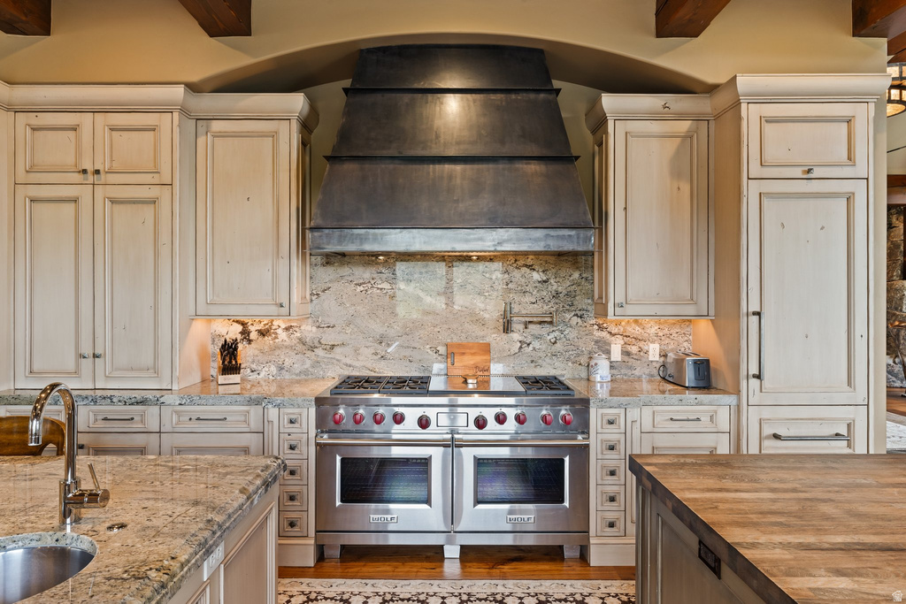 Gourmet kitchen featuring a professional-grade stainless steel range with double ovens, a matching custom metal range hood, stone slab countertops, a coordinating full-height stone backsplash, and custom wood cabinetry with distressed finish