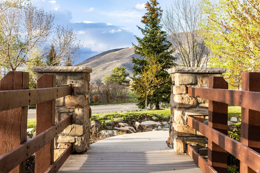 Wood-finish walkway with stone and wood-finish railing posts, framed by mature trees, with a distant mountain landscape