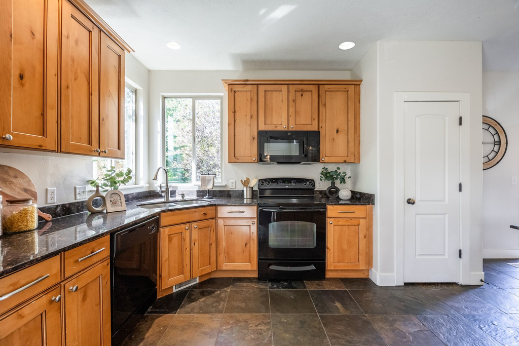 Kitchen featuring wood cabinetry, dark stone countertops, corner sink with a gooseneck faucet, black appliances, and slate tile flooring