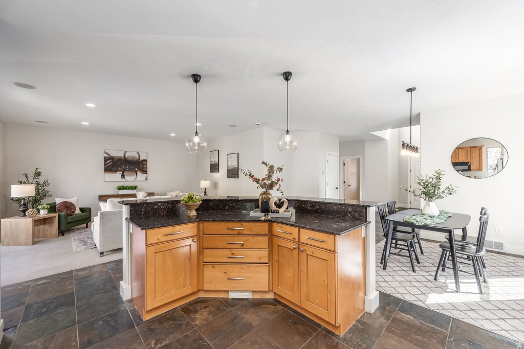 Open-concept kitchen featuring wood cabinetry, dark stone countertops, and two pendant light fixtures