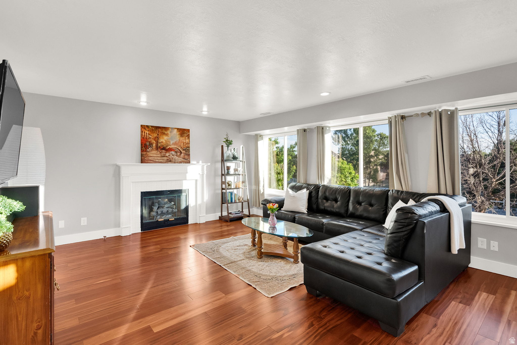 Spacious living area featuring rich wood-finish flooring, a white mantel fireplace, abundant natural light from multiple windows, and recessed lighting