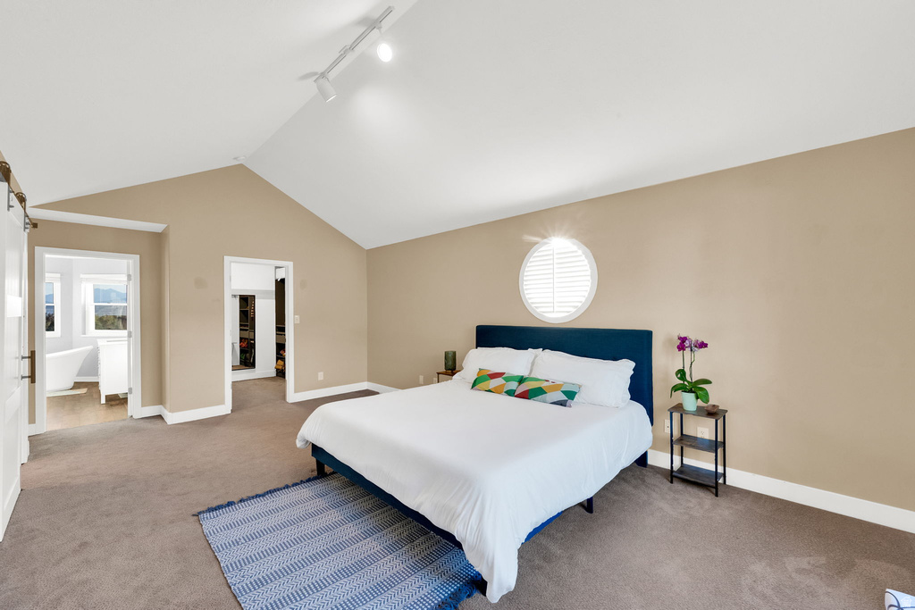 Vaulted ceilings with track lighting illuminate this carpeted room featuring a circular window with louvered shutters and a barn door entry to an additional space