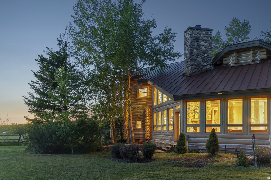 Log construction exterior featuring a stone chimney, a metal roof, multiple large windows, and surrounding mature trees