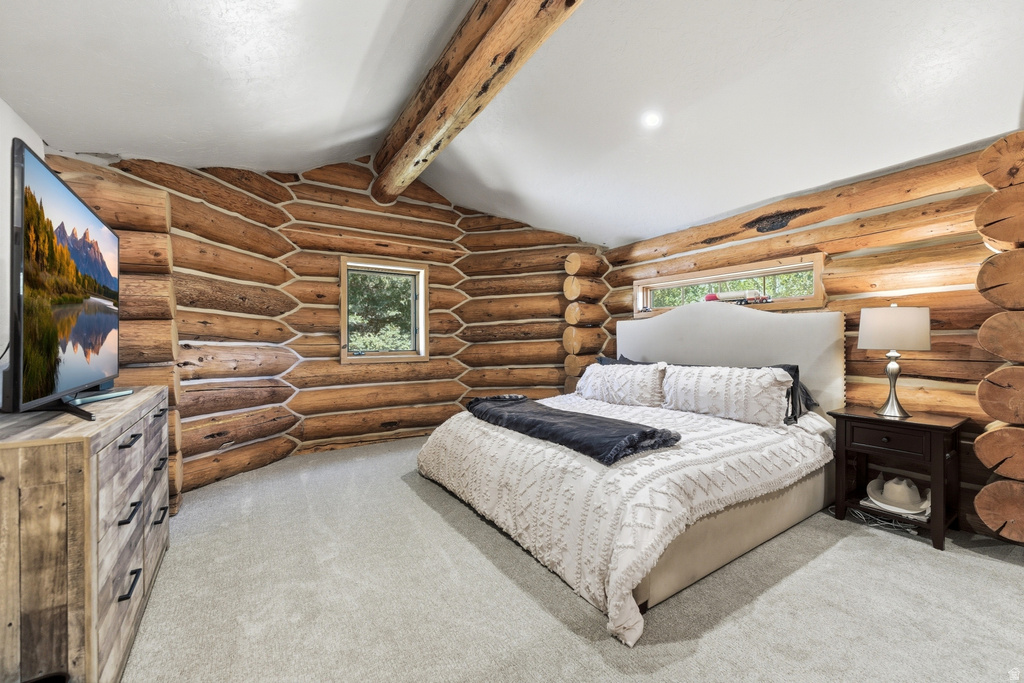 Log cabin interior featuring exposed wood log walls, a vaulted white ceiling with exposed log beams, light-colored carpeting, and two windows providing natural illumination
