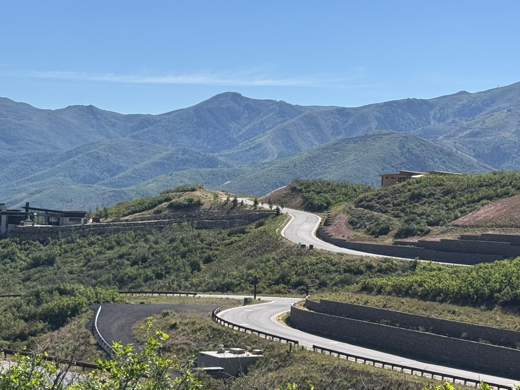 Winding asphalt road with tiered stone retaining walls, surrounded by verdant hillsides and distant mountains