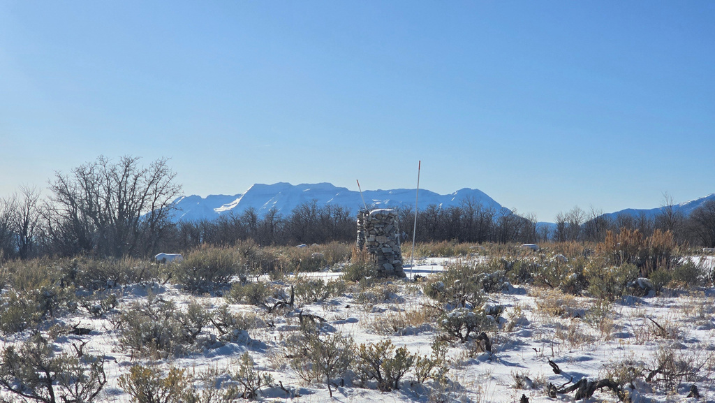 Expansive undeveloped land featuring a partial stone column, snow-dusted natural vegetation, and a clear mountain backdrop under a bright blue sky
