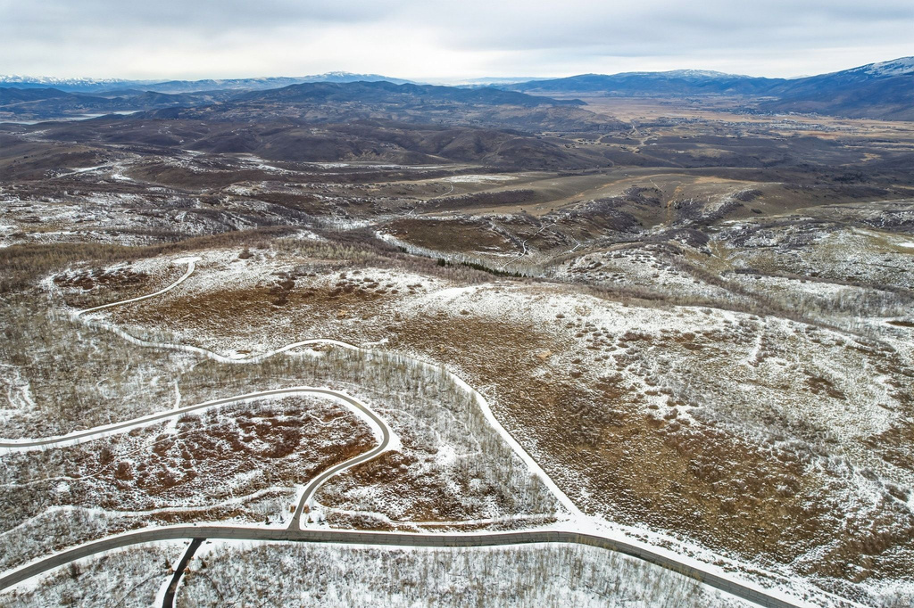 Expansive aerial landscape featuring a mountainous terrain with distant peaks, winding roads, and scattered tree cover