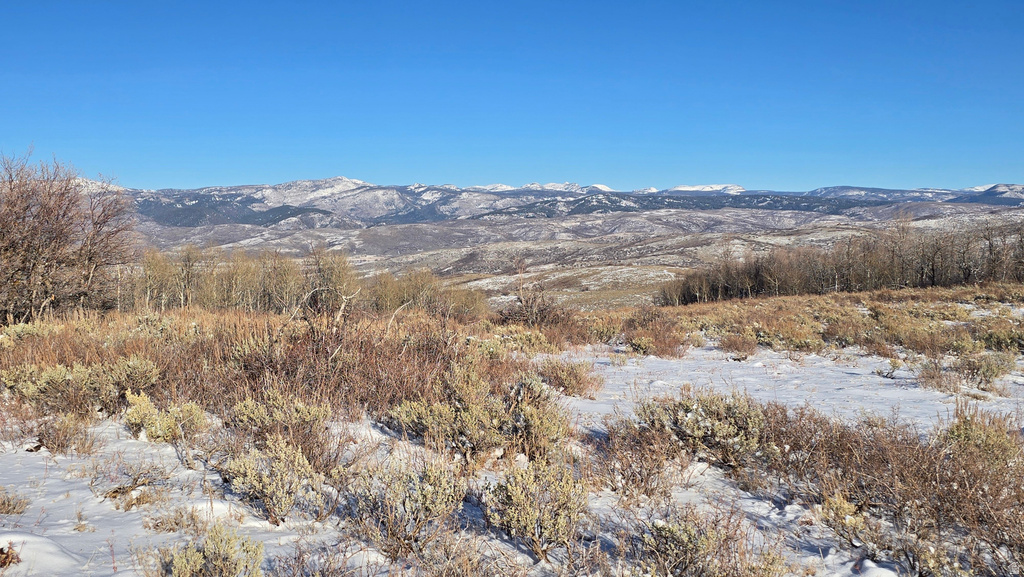 Expansive mountain landscape featuring a snow-dusted foreground, distant snow-capped peaks, and a clear blue sky