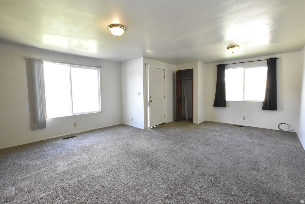 Spacious room featuring gray carpeting, white walls, two windows with vertical blinds and dark curtains, a white entry door, and a built-in closet with wood-finish trim