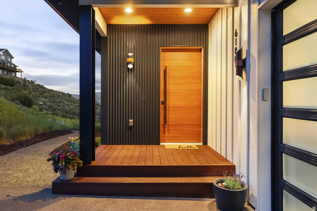 Contemporary entry featuring a wood-finish door, matching wood-finish decking, corrugated metal accent wall, vertical siding, and a modern exterior light fixture