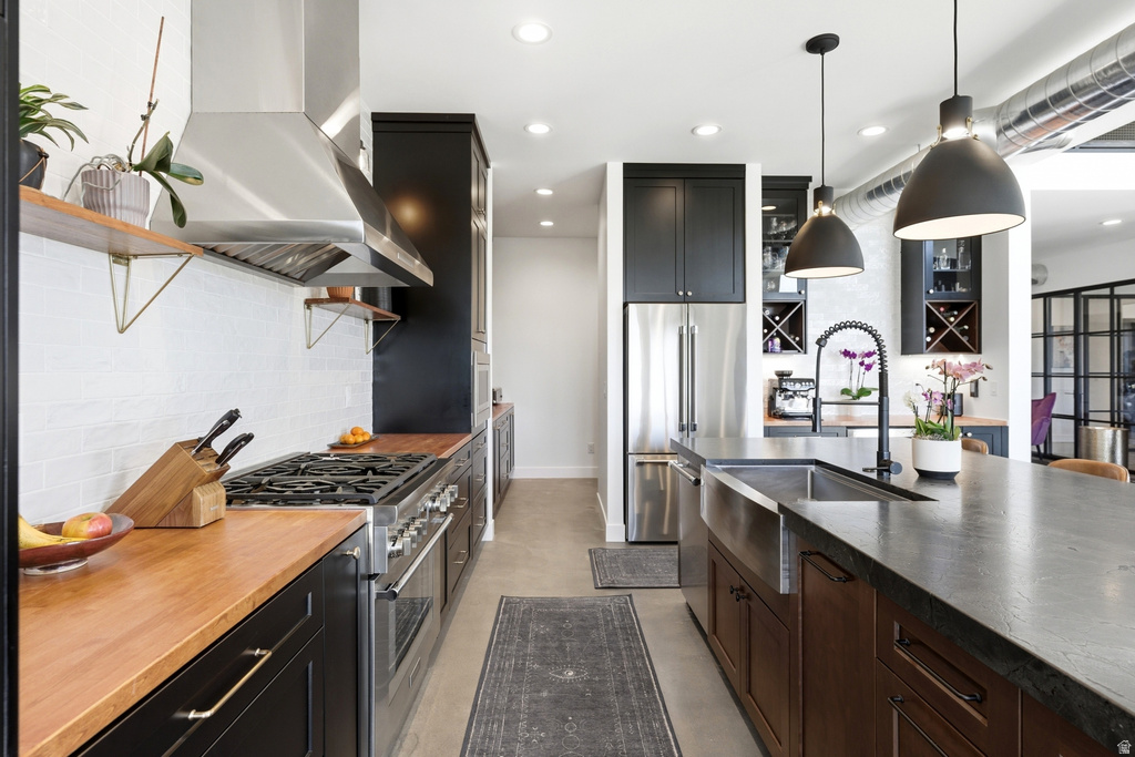 Contemporary kitchen featuring dark cabinetry, butcher block countertops, stainless steel appliances, a large island with an undermount sink, and recessed lighting
