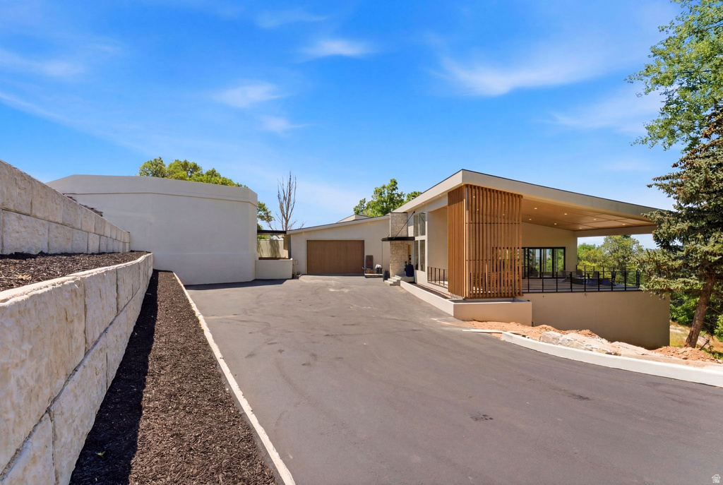 Contemporary exterior with an expansive paved driveway, layered stone retaining walls, a recessed garage with a wood-finish door, and a covered patio featuring wood-slat detailing and black railings