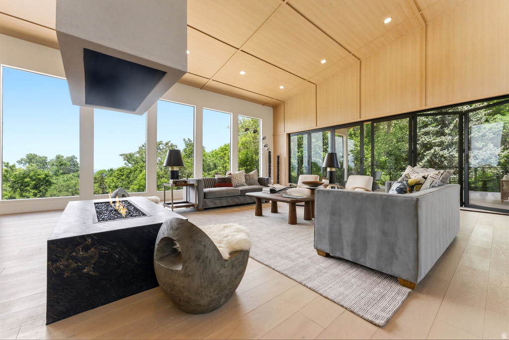 Expansive living area featuring a floating fireplace with a stone base, wood-finish flooring, a vaulted ceiling with integrated lighting, a wall of floor-to-ceiling windows, and multi-panel glass doors