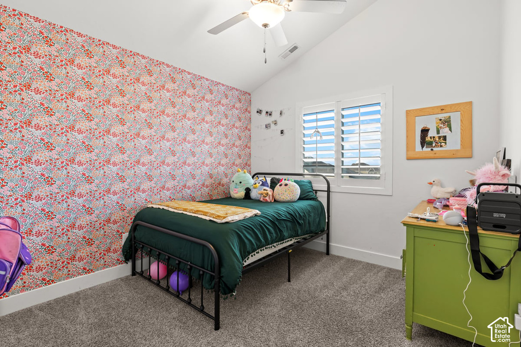 Vaulted ceiling bedroom featuring a window with plantation shutters, a patterned accent wall, plush carpet flooring, and a ceiling fan