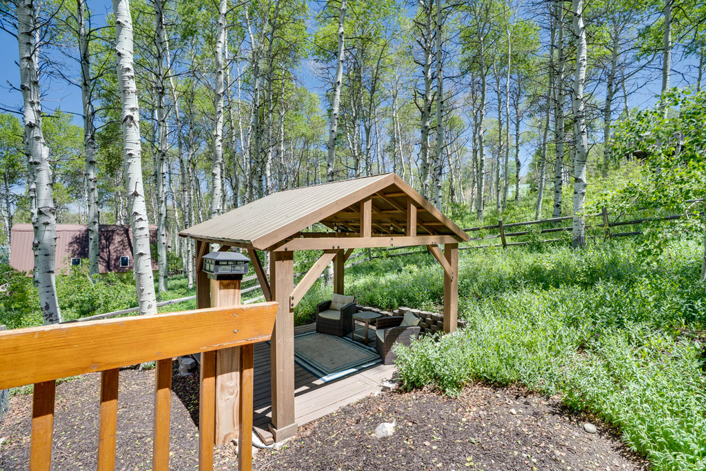 Wooden gazebo with a metal roof and integrated seating, surrounded by natural landscaping and mature trees