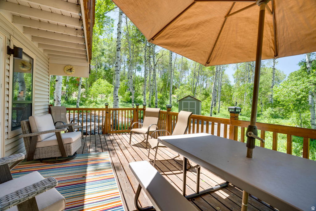 Spacious outdoor deck featuring natural wood flooring and railing, a privacy wall, a shingled shed, and surrounding mature trees