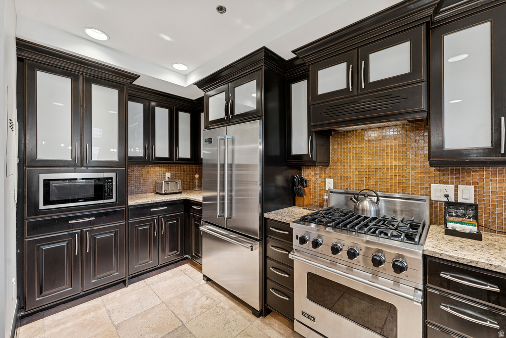Kitchen featuring dark wood cabinetry with glass-front uppers, stainless steel appliances, granite countertops, and a mosaic tile backsplash