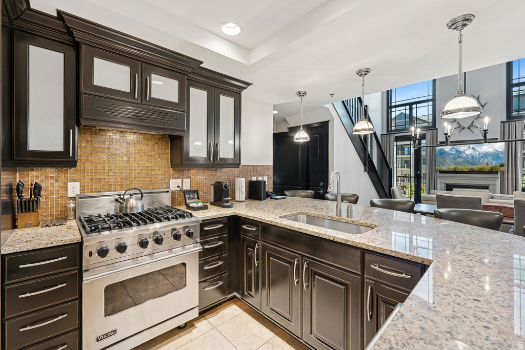Gourmet kitchen featuring a stainless steel gas range, granite countertops, dark wood-finish cabinetry with glass inserts, a mosaic tile backsplash, and a stainless steel undermount sink