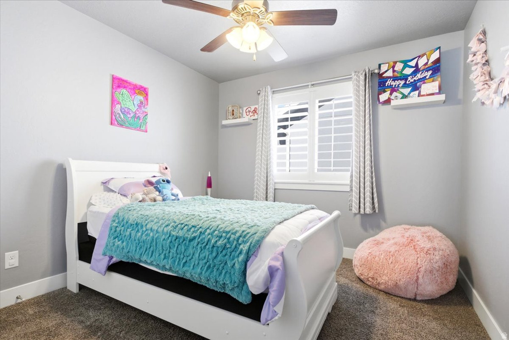 Bedroom featuring neutral gray walls, plush carpet flooring, a ceiling fan with light fixture, and a window with white louvered shutters