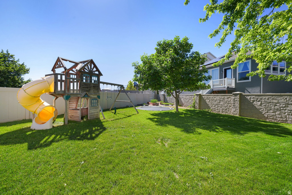 Expansive backyard featuring a manicured lawn, a wooden play structure with a yellow slide, a privacy fence, a mature shade tree, and a stone-faced retaining wall
