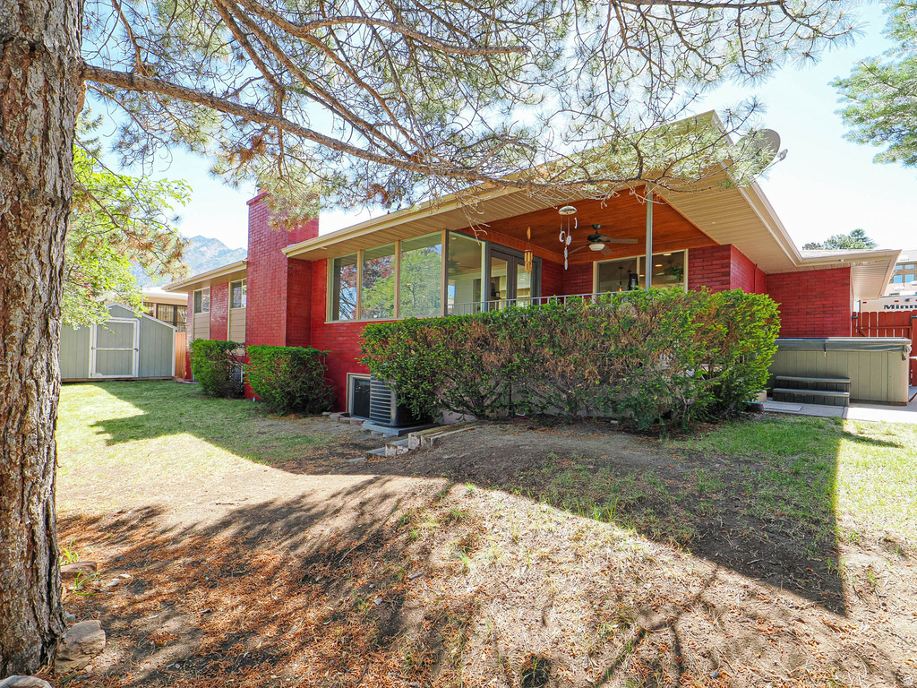 Rear exterior featuring red brick siding, a covered patio with a wood-finish ceiling, a built-in outdoor spa, a detached storage shed, and mature landscaping
