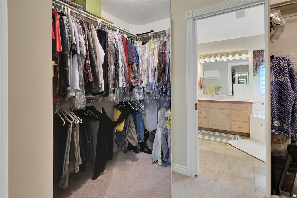 Expansive walk-in closet with carpet flooring and multi-level hanging rods, offering direct access to a bathroom featuring a light wood-finish vanity with dual sinks, a large framed mirror, and a white soaking tub