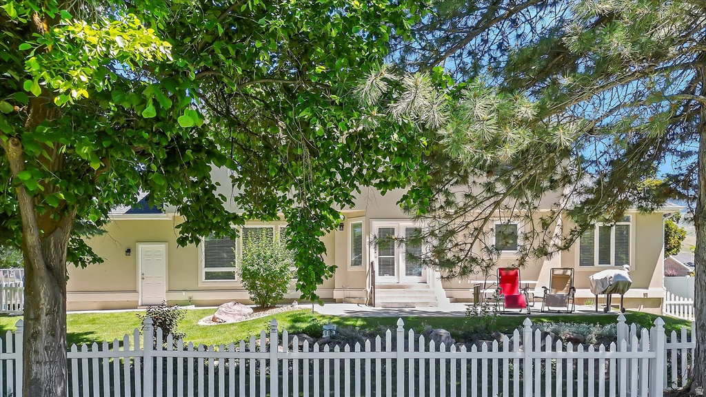 Rear exterior featuring a white picket fence, a manicured lawn, a concrete patio, and mature trees