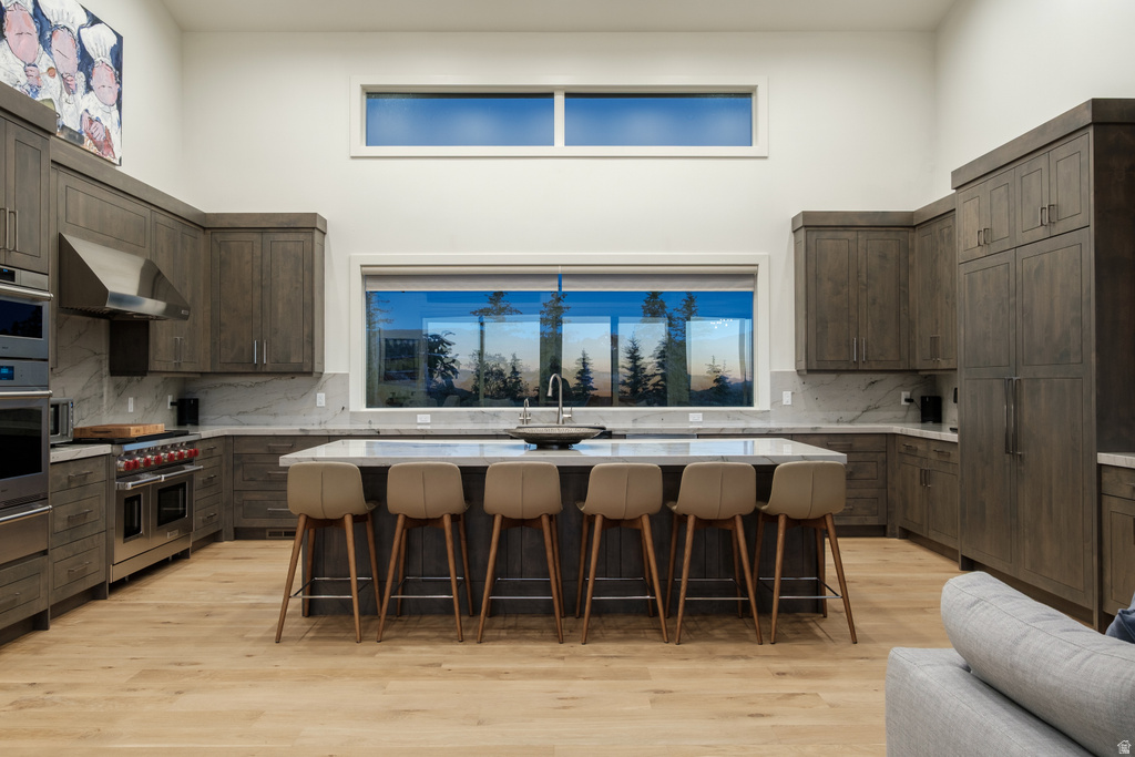 Expansive kitchen featuring wood-finish cabinetry, stainless steel appliances, a large center island with counter seating, a stone countertop, and a wide window overlooking a treed landscape