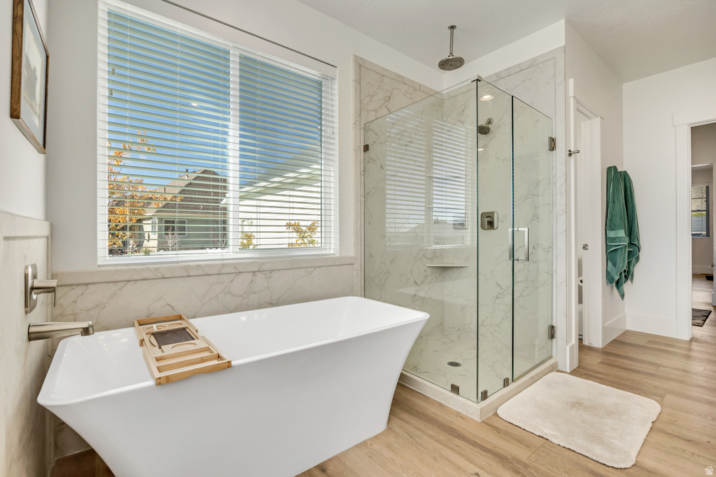 Freestanding soaking tub with chrome fixtures, glass-enclosed shower with a rainfall showerhead, large window with blinds, wood-finish flooring, and marble-look wall tile accents