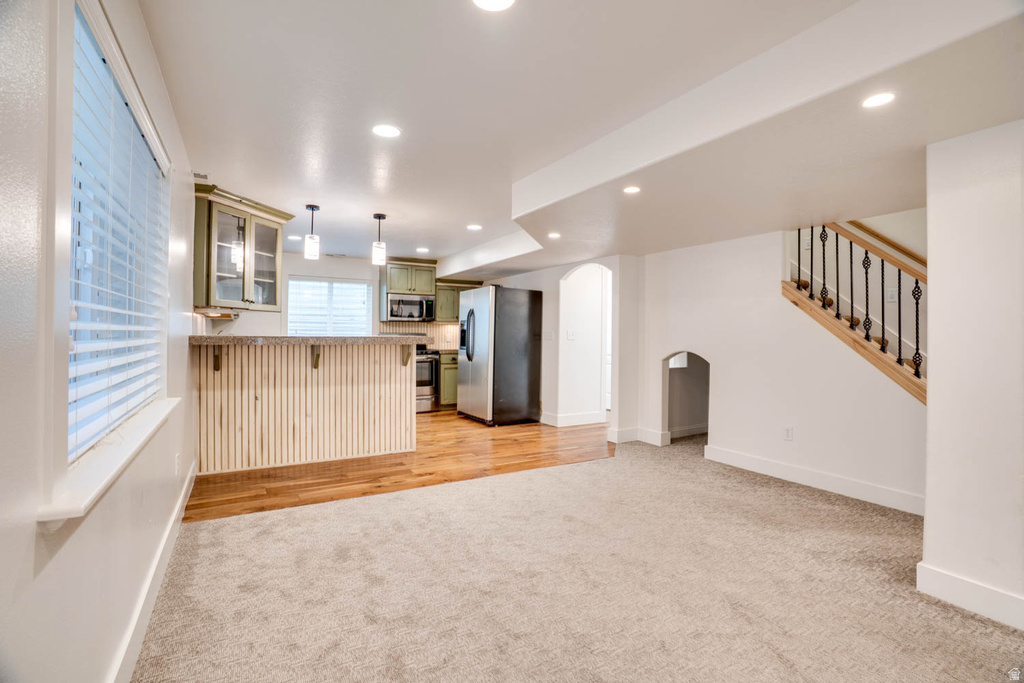 Open-concept living space featuring neutral carpeting, wood-finish flooring, a kitchen area with light green cabinetry and pendant lighting, and a staircase with wrought iron balusters