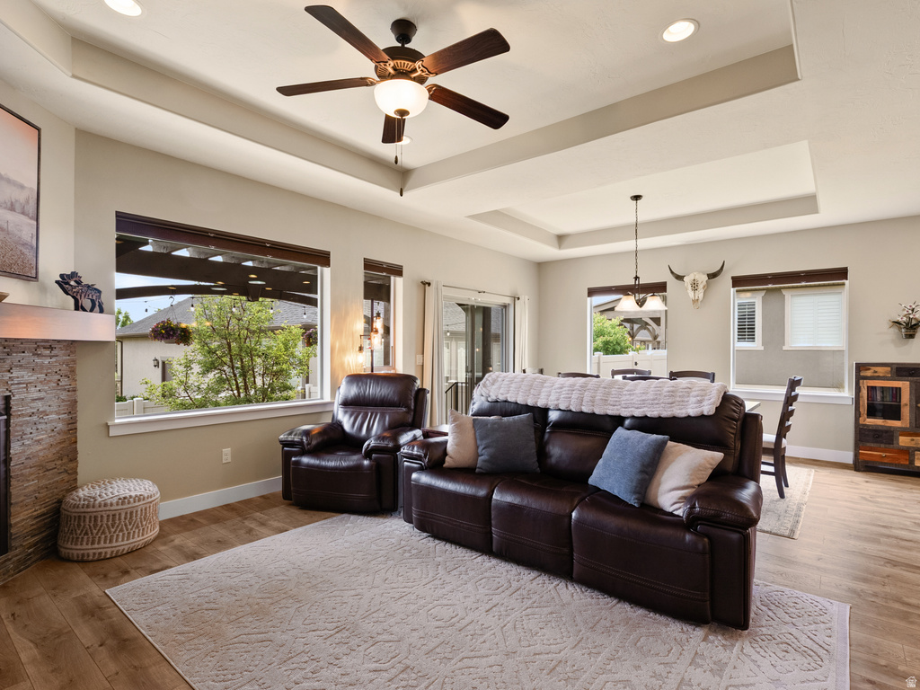 Spacious living area featuring wood-finish flooring, a tray ceiling with recessed lighting and a ceiling fan, and a stacked stone fireplace
