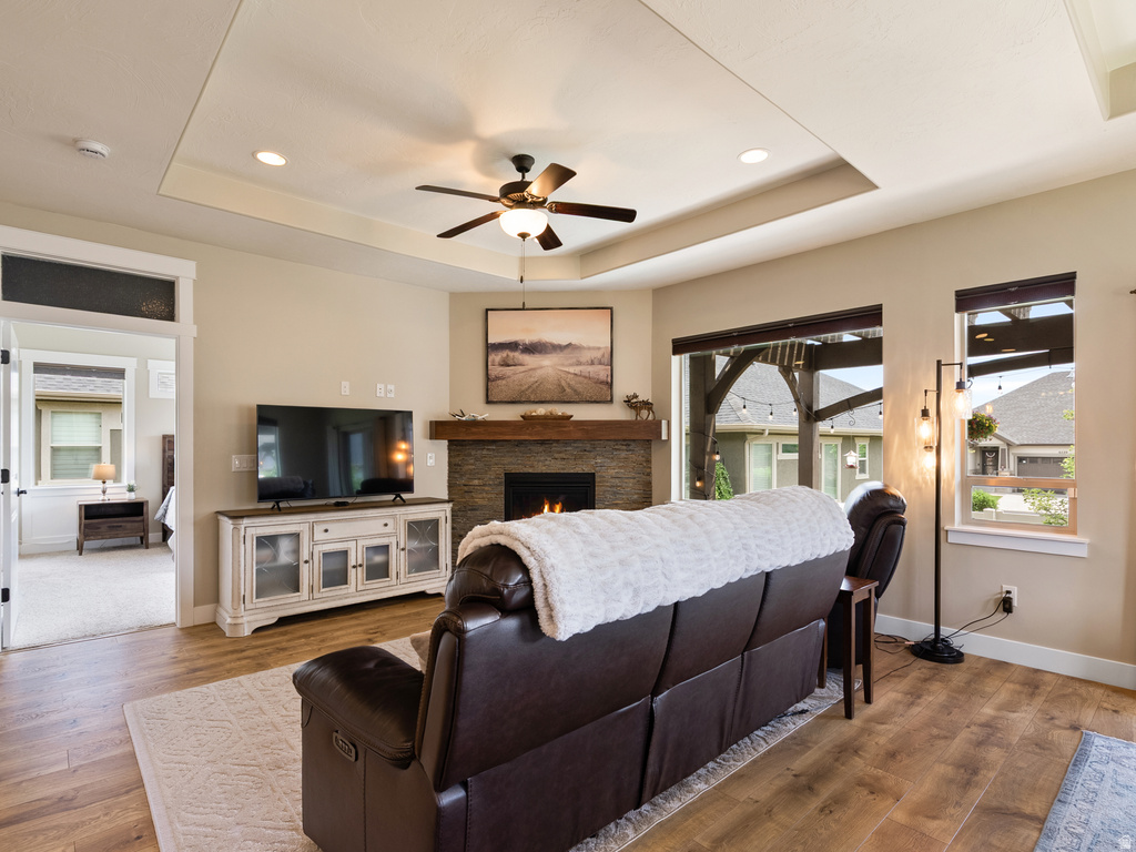 Living area featuring wood-finish flooring, a stone-front fireplace with a wood mantle, and a tray ceiling with recessed lighting and a ceiling fan