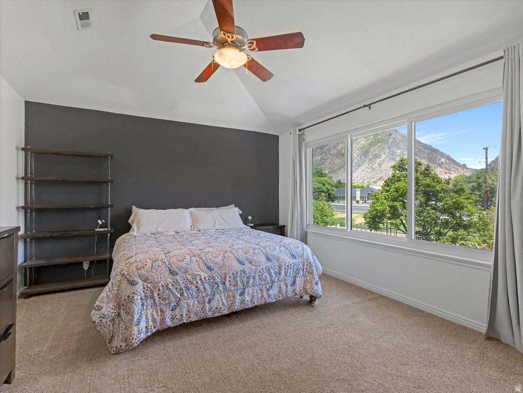 Bedroom featuring vaulted ceiling, ceiling fan, carpet flooring, a dark accent wall, and multiple windows with a mountain backdrop