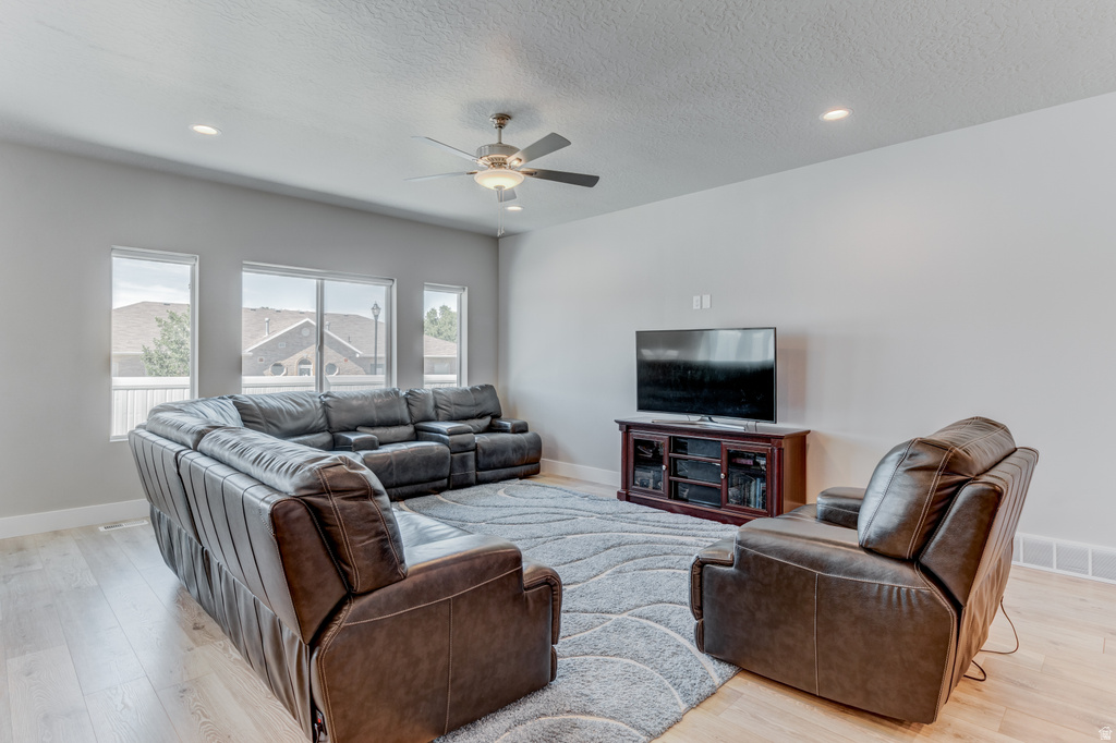Spacious living area featuring light wood-finish flooring, a contemporary ceiling fan with integrated lighting, recessed ceiling lights, and multiple windows