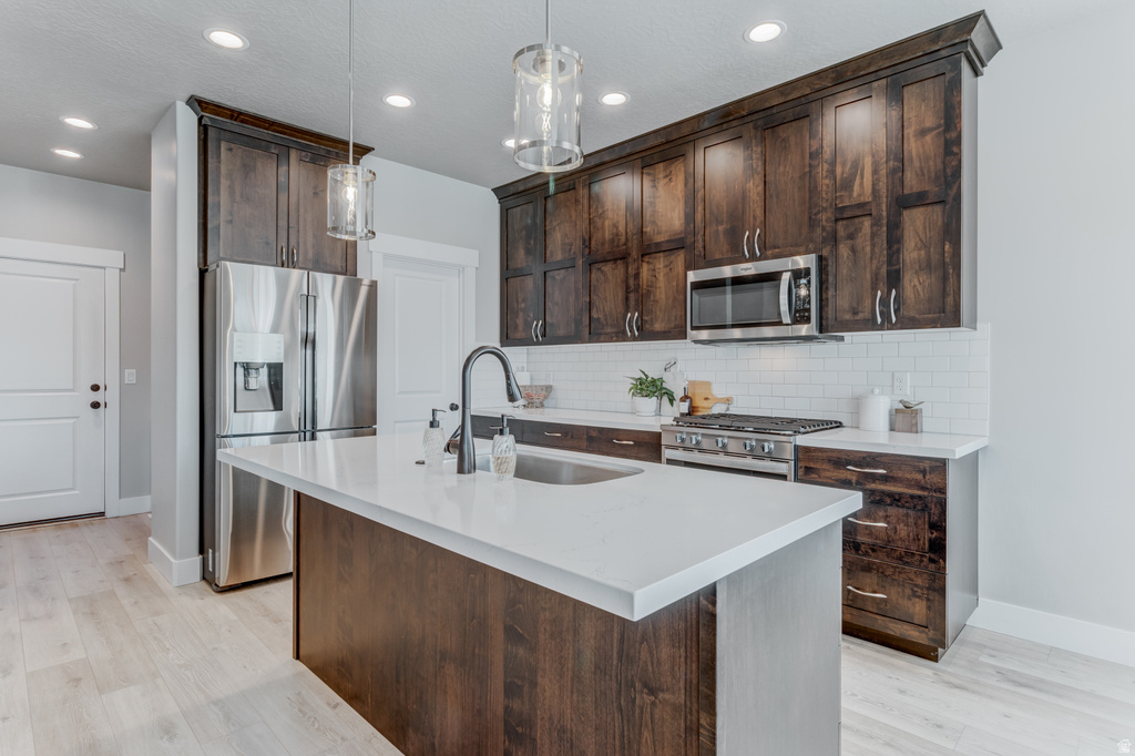 Kitchen featuring a central island with an undermount sink, dark wood cabinetry, stainless steel appliances, white subway tile backsplash, and light wood-finish flooring