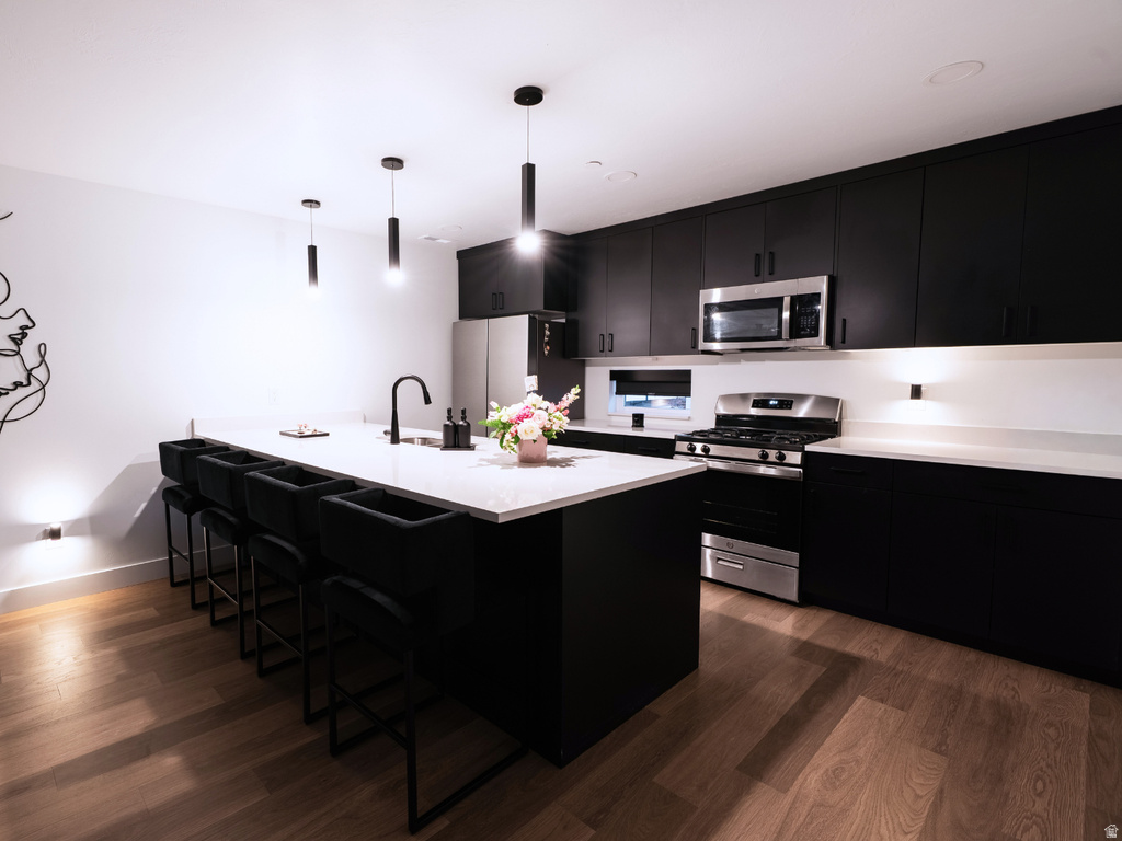 Modern kitchen featuring dark cabinetry, light countertops, wood-finish flooring, stainless steel appliances, and a central island with pendant lighting