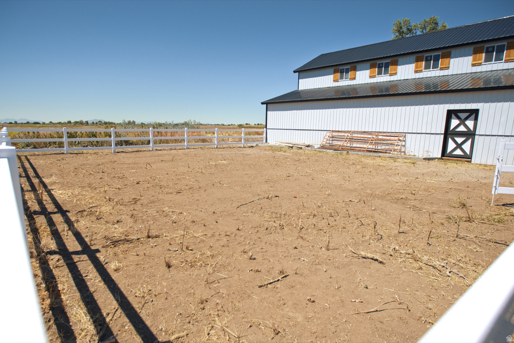 Expansive outdoor area with a white two-rail fence, a metal-roofed barn-style structure featuring white vertical siding, multiple windows with natural wood shutters, and a black and white crossbuck door