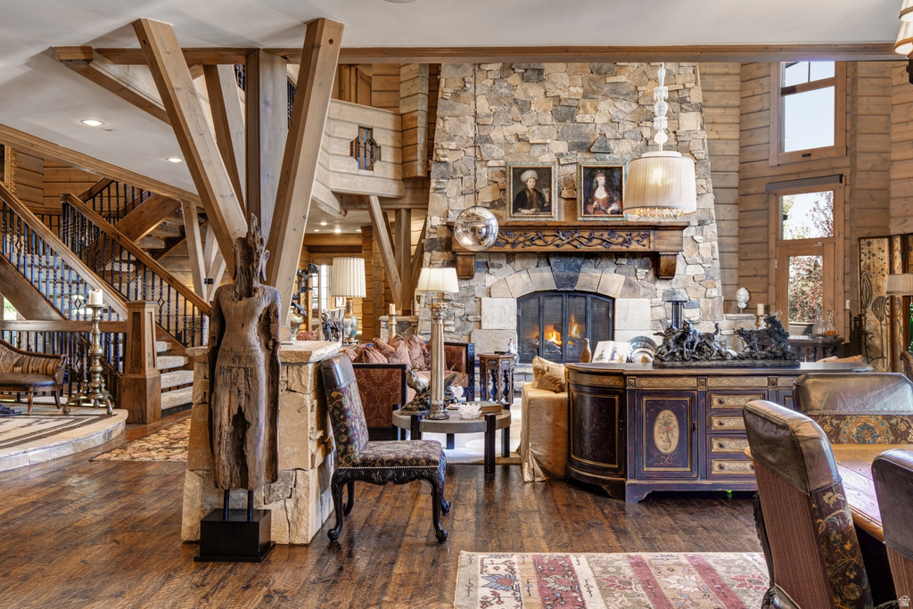 Great room featuring a floor-to-ceiling stone fireplace with a carved wood mantel, exposed timber beams, wood-finish flooring, and a grand staircase with dark iron balusters