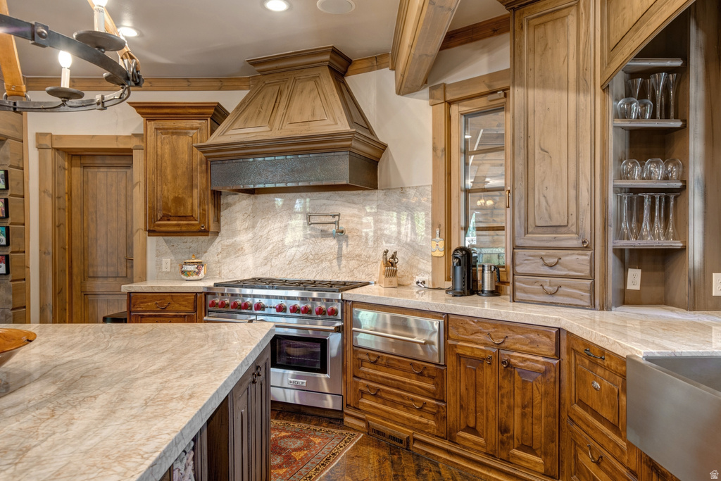 Gourmet kitchen featuring a built-in stainless steel range with a custom wood hood, light-toned stone countertops, a full stone backsplash, and wood cabinetry throughout