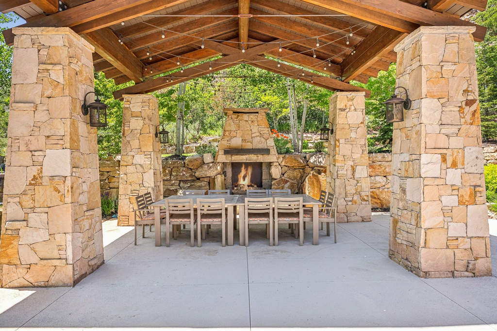 Expansive outdoor patio featuring a stone fireplace, wood-beamed ceiling, integrated lighting, stone support pillars, and concrete flooring