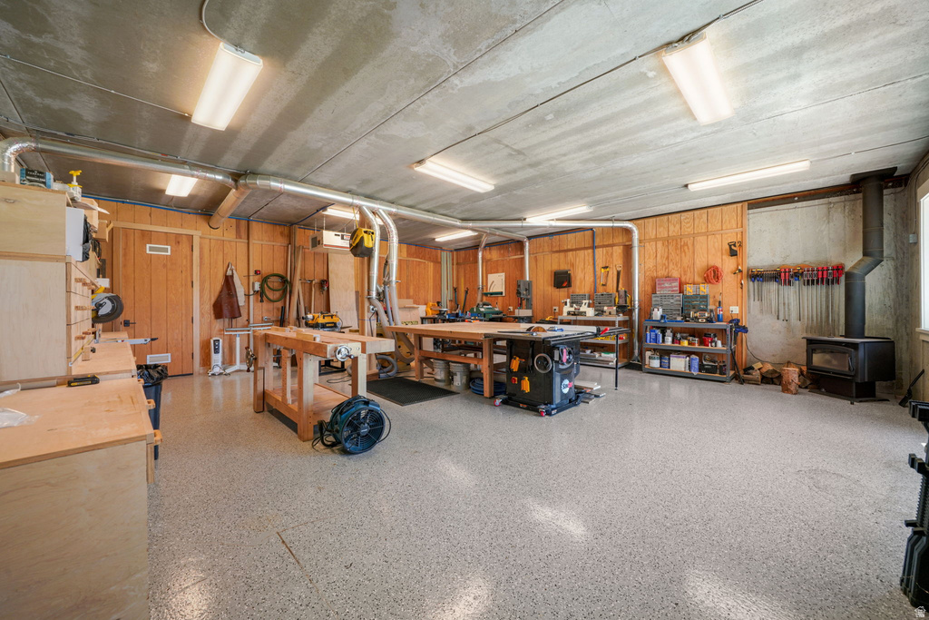 Spacious workshop featuring wood paneling, a wood-burning stove, exposed ceiling ductwork, multiple fluorescent light fixtures, and a durable epoxy-coated concrete floor
