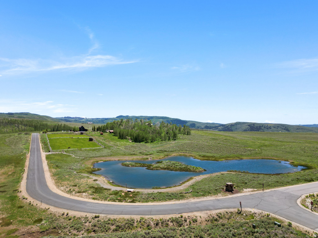 Expansive landscape featuring an asphalt road, a natural pond, and distant structures surrounded by rolling green hills and scattered trees